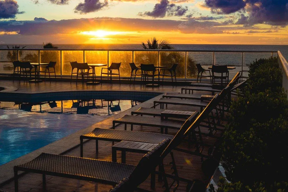 Piscina do rooftop do Hotel Ponta Verde Praia do Francês com o sol baixo no horizonte do mar, mesas junto ao parapeito de vidro e espreguiçadeiras na borda