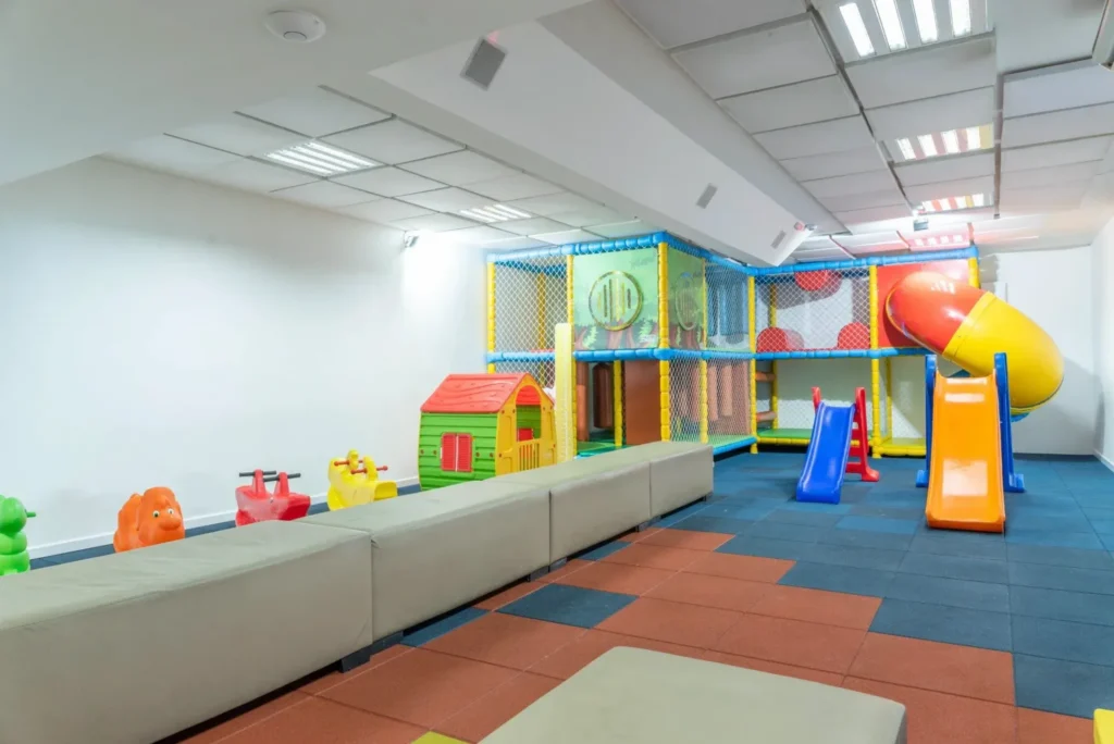 Sala de recreação infantil coberta, com casinha, escorregadores e brinquedão de plástico sobre piso emborrachado